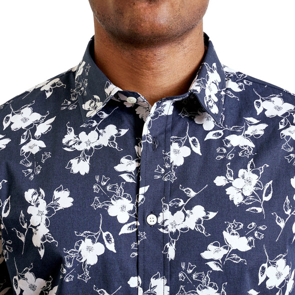 close up view of the pointed collar and neck area of cotton fabric button up shirt in a white floral pattern on navy ground. Worn by a black male model. The top buttons are undone